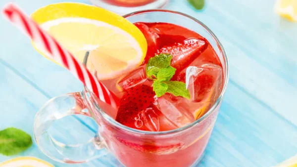 A glass of strawberry lemonade sitting on a blue table with a straw and lemon on the rim